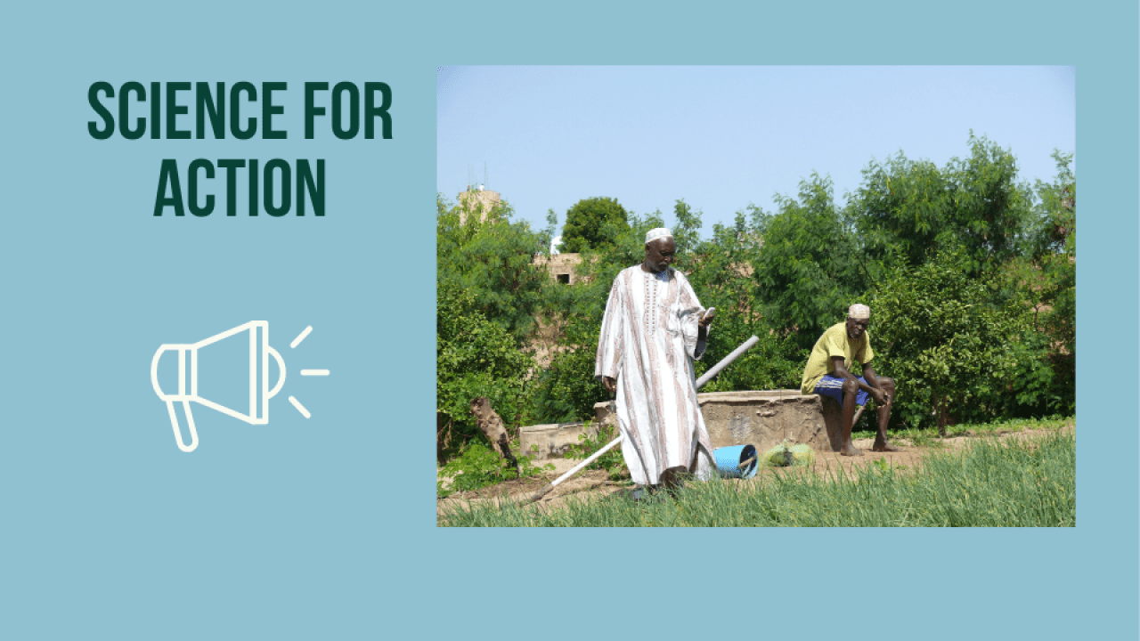 Two men one standing holding a phone and one sitting on a well
